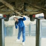 Mechanik w niebieskim kombinezonie ogląda od spodu uniesiony samochód